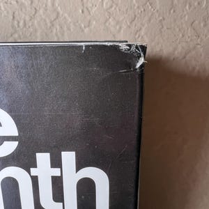 May include: Close-up of a book's corner with the title partially visible. The book has a black cover with white lettering. The corner shows wear and tear, with a small section torn away. The background is a neutral beige wall.