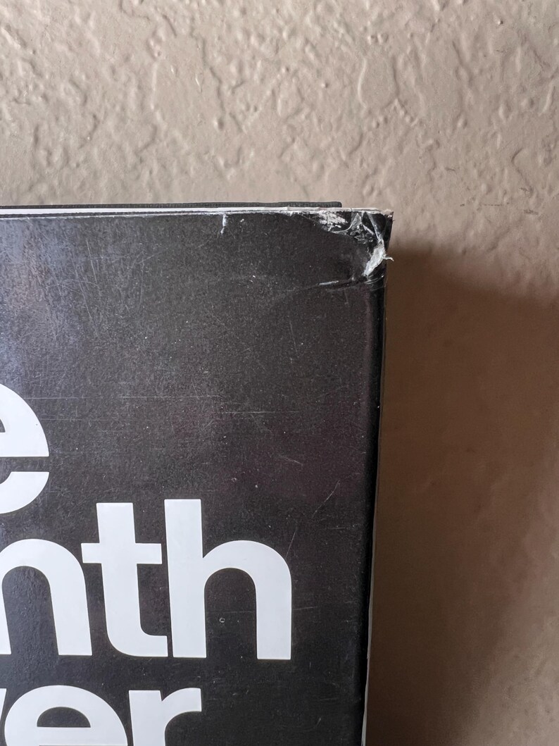 May include: Close-up of a book's corner with the title partially visible. The book has a black cover with white lettering. The corner shows wear and tear, with a small section torn away. The background is a neutral beige wall.