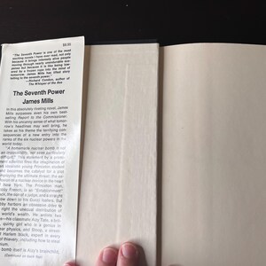 May include: Open book with the title "The Seventh Power" by James Mills. The book's spine and pages are visible, with text on the inside cover and a price tag. The book is open, revealing the inside pages.