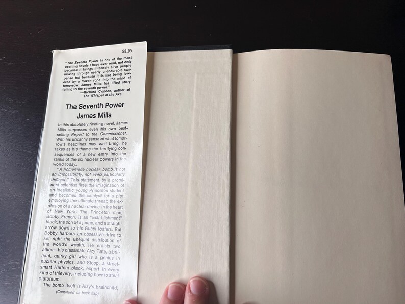 May include: Open book with the title "The Seventh Power" by James Mills. The book's spine and pages are visible, with text on the inside cover and a price tag. The book is open, revealing the inside pages.