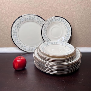 May include: A stack of 12 white ceramic plates with a silver rim and a floral design. The plates are arranged in a stack with the largest plate on the bottom and the smallest plate on top. There is a red apple in the foreground.