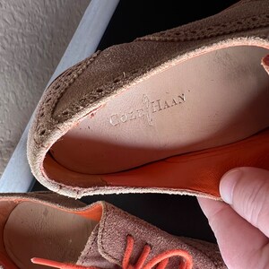 Puede incluir: Primer plano de un par de zapatos de ante marr&oacute;n con cordones naranjas. Los zapatos tienen un dise&ntilde;o de brogue y el nombre de la marca "Cole Haan" es visible en la plantilla.