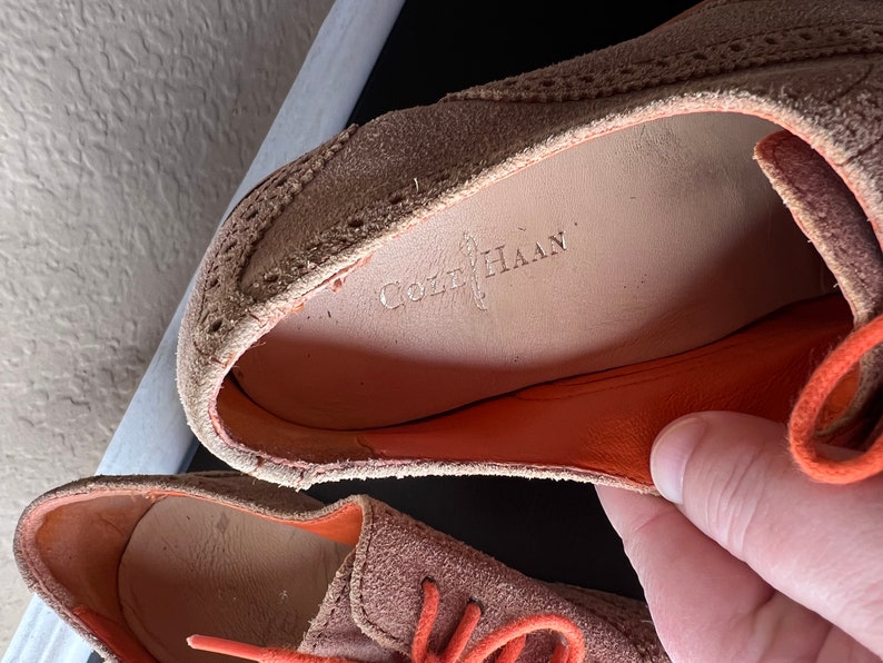 Puede incluir: Primer plano de un par de zapatos de ante marr&oacute;n con cordones naranjas. Los zapatos tienen un dise&ntilde;o de brogue y el nombre de la marca "Cole Haan" es visible en la plantilla.