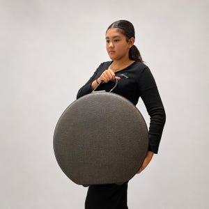 May include: A large, circular, dark gray suitcase with a textured fabric finish and a single top handle. The case features a minimalist, rounded design with a dark zipper closure running along the perimeter, providing a structured and portable storage solution.