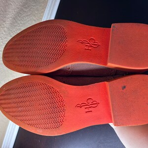 Puede incluir: Primer plano de las suelas naranjas de un par de zapatos de ante marr&oacute;n. Las suelas tienen un patr&oacute;n texturizado y el nombre de la marca "CPH" est&aacute; impreso en la parte inferior.