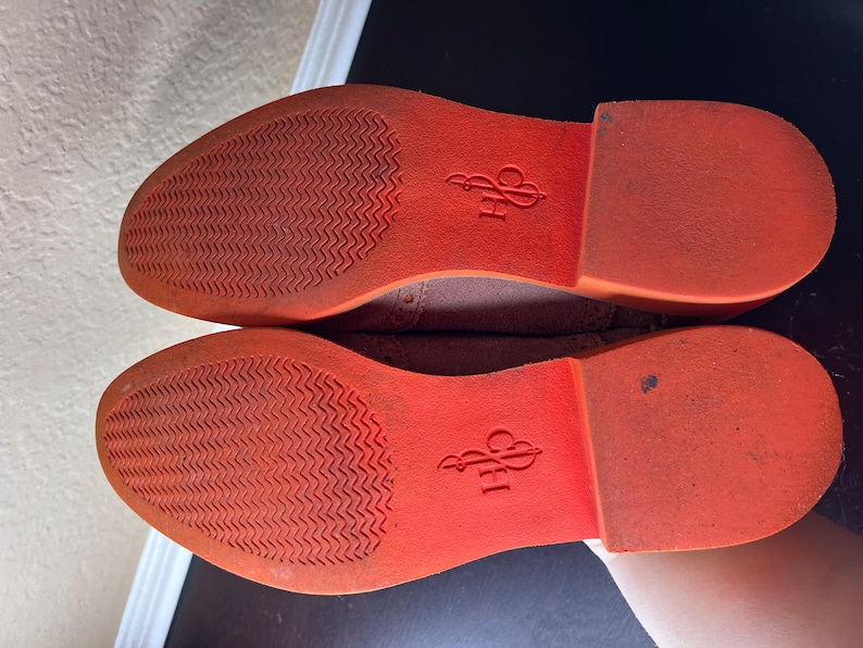 Puede incluir: Primer plano de las suelas naranjas de un par de zapatos de ante marr&oacute;n. Las suelas tienen un patr&oacute;n texturizado y el nombre de la marca "CPH" est&aacute; impreso en la parte inferior.