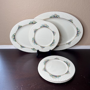 May include: Set of four white oval and round ceramic plates with a floral border design in shades of green, purple, and yellow. The plates have a gold rim and a fluted edge.