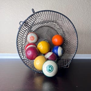Könnte beinhalten: Eine Sammlung von Billardkugeln in einem Drahtgitterkorb. Die Kugeln sind in verschiedenen Farben, darunter Rot, Gelb, Orange und Blau, mit Zahlen auf einigen sichtbar. Der Korb ist schwarz und hat einen Griff.
