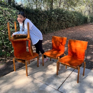 May include: Three orange upholstered chairs with wooden frames. The chairs are arranged in a row, with the middle chair slightly offset. A person is standing behind the chairs, leaning against the back of the chair on the left.
