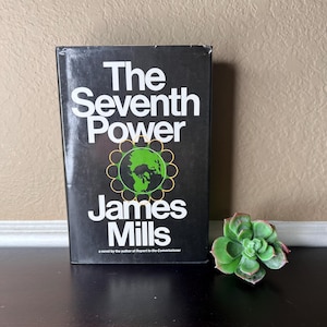 May include: A hardcover book titled "The Seventh Power" by James Mills, with a black cover and white text. The cover features a green and gold graphic of the Earth. A succulent plant is next to the book.
