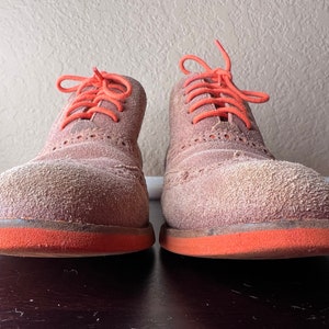 Puede incluir: Un par de zapatos de ante marr&oacute;n con cordones naranjas y ribete naranja alrededor de las suelas. Los zapatos est&aacute;n polvorientos y parecen usados.