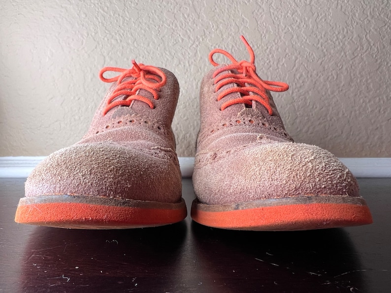 Puede incluir: Un par de zapatos de ante marr&oacute;n con cordones naranjas y ribete naranja alrededor de las suelas. Los zapatos est&aacute;n polvorientos y parecen usados.