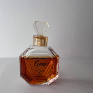May include: A clear glass perfume bottle with an octagonal shape containing amber liquid. The bottle features a gold-toned collar and a faceted, diamond-shaped clear stopper. The text reads: eau de toilette Gem de Van Cleef & Arpels Paris.