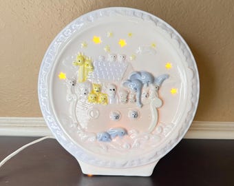 May include: A white ceramic night light featuring a relief of Noah's Ark with various animals. The design includes a giraffe, elephant, and other pairs of animals. The light emits a soft glow, creating a warm ambiance. The night light is round with a decorative edge.