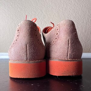 Puede incluir: Un par de zapatos de ante marr&oacute;n con suelas naranjas. Los zapatos tienen un dise&ntilde;o de brogue y est&aacute;n atados.