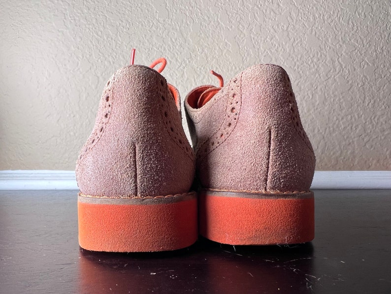 Puede incluir: Un par de zapatos de ante marr&oacute;n con suelas naranjas. Los zapatos tienen un dise&ntilde;o de brogue y est&aacute;n atados.