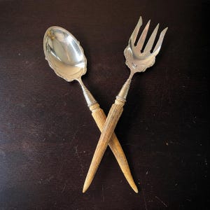 May include: Silver serving spoon and fork crossed on a dark brown surface. The spoon and fork have ornate silver heads and light brown, textured handles. The spoon has a scalloped edge.