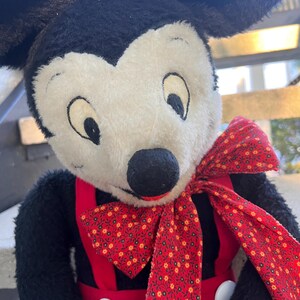 May include: A plush Mickey Mouse doll with black fur, a white face, and large black ears. He wears red overalls with white buttons and a red bow tie patterned with small yellow flowers. The doll has a classic, vintage appearance.