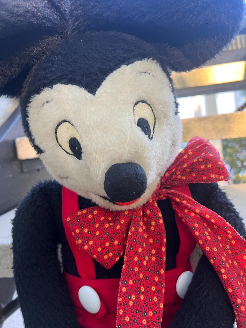 May include: A plush Mickey Mouse doll with black fur, a white face, and large black ears. He wears red overalls with white buttons and a red bow tie patterned with small yellow flowers. The doll has a classic, vintage appearance.