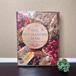 Peut inclure: Un livre vintage intitulé "The Needlework Book" de Wanda Passadore. La couverture présente un collage coloré de fournitures de broderie, notamment des cerceaux à broder, des chutes de tissu et de la laine.