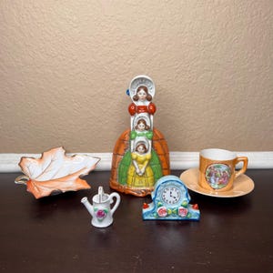 May include: A set of vintage porcelain figurines, including a three-tiered doll figurine, a small watering can, a leaf-shaped dish, and a clock figurine with floral accents. The figurines are painted in bright colors and feature a whimsical, fairytale-like theme.