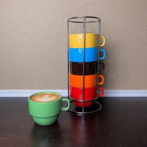 May include: A stack of colorful coffee cups in yellow, blue, brown, orange, and red, held in a chrome rack. A green coffee cup with a latte sits in front of the stack. The latte has a decorative design.