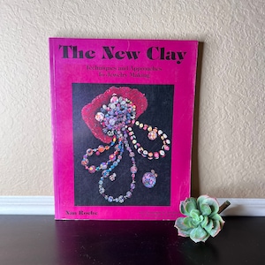 May include: A pink book titled "The New Clay: Techniques and Approaches to Jewellery Making" by Nan Roche. The cover features a colourful illustration of jewellery made with clay.