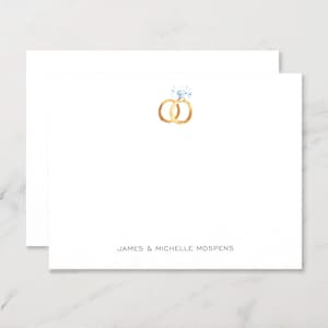 May include: White stationery with a watercolor illustration of two gold wedding rings, one with a diamond. The names "JAMES & MICHELLE MOSPENS" are printed in a simple font at the bottom of the card.