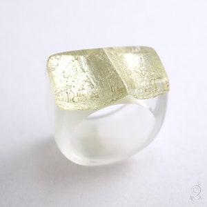 Può includere: Un anello in resina bianca con un design a foglia d'oro sulla parte superiore. L'anello è spesso e ha una fascia larga.