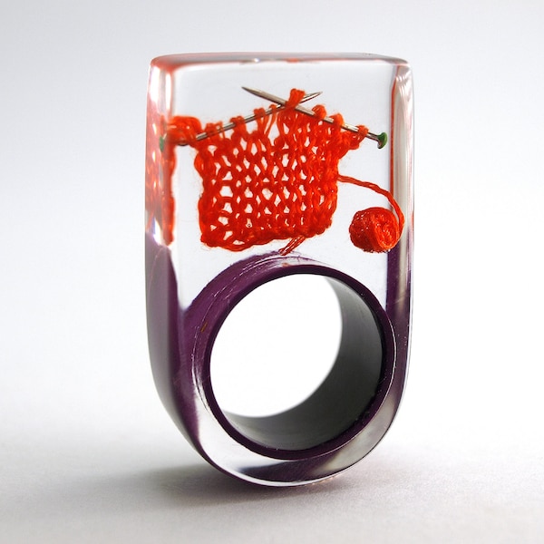 Strickzeug – Außergewöhnlicher Strick-Ring mit orange-rot gestrickter Wolle und Stricknadeln auf lila Ring in Gießharz