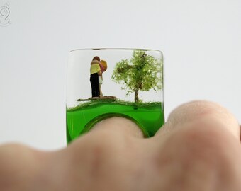 Chaleur du cœur - Bague des amoureux au paradis avec des mini-figurines qui s'embrassent à côté d'un arbre et un serpent sur une bague verte en résine coulée pour les amoureux