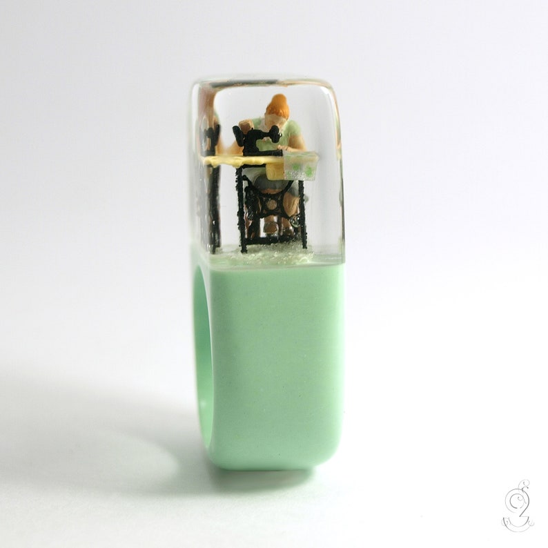 Op de afbeelding: Een unieke ring met een miniatuursc&egrave;ne van een persoon die naait op een vintage naaimachine. De ring heeft een lichtgroene band en een transparante, rechthoekige bovenkant. De miniatuursc&egrave;ne staat tegen een witte achtergrond.