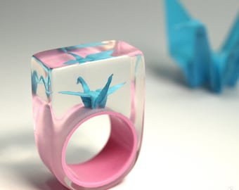 May include: A clear resin ring featuring an embedded light blue paper crane. The ring has a thick, rectangular top section and a solid pink inner band. A separate light blue paper crane is positioned out of focus in the background.