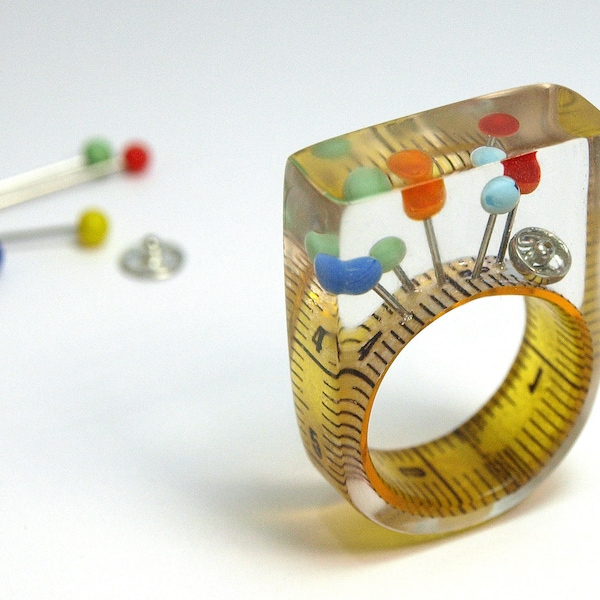 Nähkästchen – Ungewöhnlicher Nähutensilien-Ring mit Maßband, bunten Stecknadeln und Druckknopf auf gelbem Ring in Gießharz