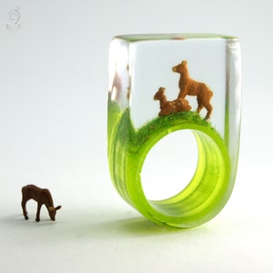Puede incluir: Un anillo de resina transparente con una base verde y dos figuritas de ciervos en miniatura en su interior. Los ciervos son marrones y están de pie en una zona verde cubierta de hierba.