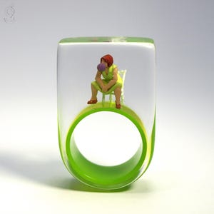 Può includere: Un anello unico con una parte superiore trasparente e rettangolare e una fascia verde brillante. All'interno, una figura in miniatura di una persona con un vestito verde seduta su una sedia bianca, che soffia una bolla. Il design dell'anello è artistico e accattivante.