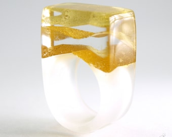 May include: A ring crafted from clear resin featuring a rectangular top section with embedded layers of shimmering gold-colored flakes. The lower portion of the band is made of a solid, opaque white material, creating a distinct contrast with the transparent upper part.