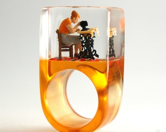 May include: A clear resin ring featuring an embedded miniature scene of a person with reddish hair wearing an orange top and grey skirt, sitting on a brown chair at a black vintage sewing machine on a colorful patterned rug base.