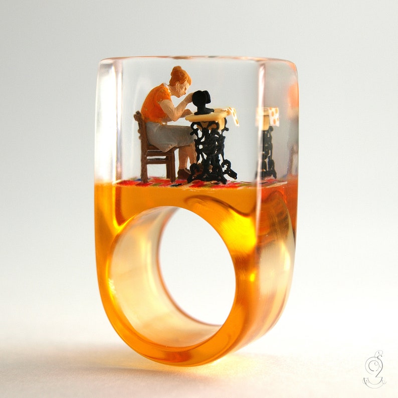 Op de afbeelding: Een unieke harsring met een miniatuursc&egrave;ne van een persoon die naait op een vintage naaimachine. De ring is transparant met een amberkleurige basis en de miniatuursc&egrave;ne staat op een kleurrijk tapijt.