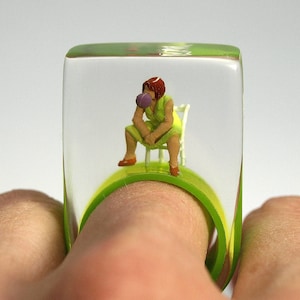 Bubble Gum Queen – Cheeky figures ring with a woman sitting on a chair, making a chewing gum bubble, on a colorful ring made of resin