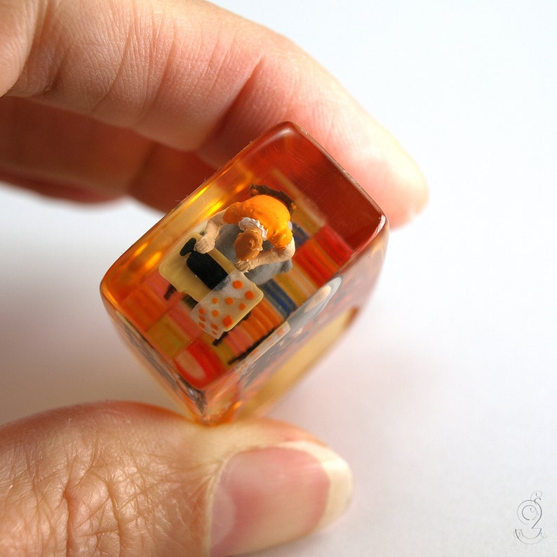 Op de afbeelding: Een miniatuur diorama ingekapseld in een heldere, amberkleurige harskubus. De sc&egrave;ne toont een klein figuur in een oranje shirt en witte schort, zittend op een kleurrijk gestreept tapijt. De kubus wordt tussen vingers gehouden.