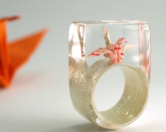May include: A clear resin ring featuring a miniature origami crane with red and white striped patterns embedded inside. The ring has a thick, rectangular profile with a smooth, transparent finish and a textured, metallic-toned inner band structure.
