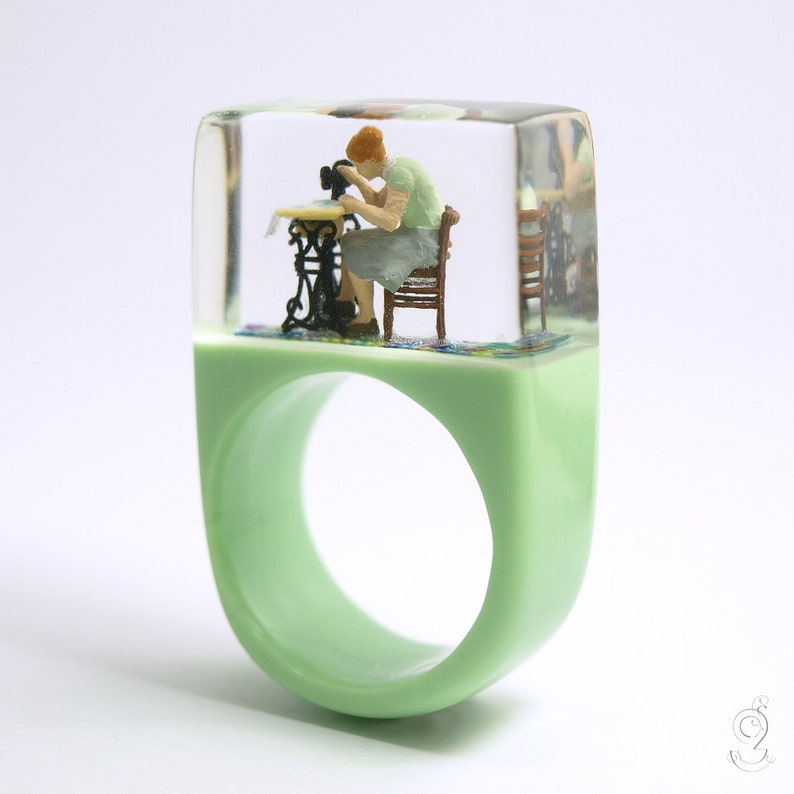 Op de afbeelding: Een lichtgroene harsring met een rechthoekige bovenkant. Binnenin de heldere hars bevindt zich een miniatuursc&egrave;ne van een persoon die naait aan een vintage naaimachine. De ring heeft een gladde, afgeronde band.