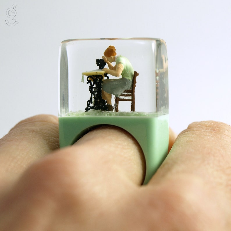 Op de afbeelding: Een unieke ring met een miniatuursc&egrave;ne van een persoon die naait op een vintage naaimachine. De heldere harskubus omsluit de kleine figuren en machine, geplaatst op een lichtgroene ringbasis. De ring wordt door een hand vastgehouden.
