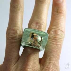 Op de afbeelding: Een unieke, vierkante ring met een heldere, lichtgroene harszetting. Binnenin de hars bevindt zich een miniatuursc&egrave;ne van een persoon die aan een bureau werkt. De ring wordt aan een vinger gedragen.