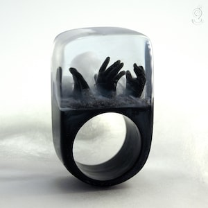 May include: A unique, handcrafted ring featuring a black band and a clear resin top. Inside the resin, black hands appear to be reaching upwards from a sandy base. The ring has a modern, artistic design.
