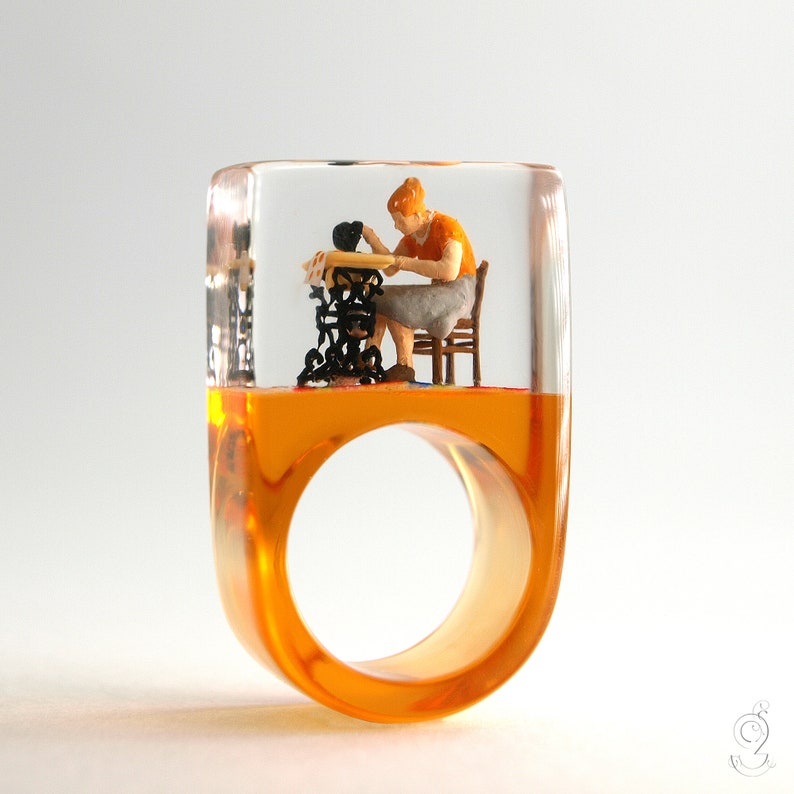 Op de afbeelding: Een unieke ring met een heldere, rechthoekige bovenkant en een oranje band. Binnenin de heldere bovenkant zit een miniatuurfiguur van een persoon die aan een naaimachine zit. Het ontwerp van de ring is artistiek en opvallend.