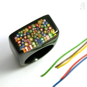 Puede incluir: Un anillo de resina negra con una parte superior rectangular, que muestra un mosaico de cables coloridos cortados transversalmente. Los cables son de varios colores, incluyendo rojo, amarillo, verde y azul. Varios cables sueltos en colores a juego están en el fondo.