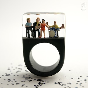 Könnte beinhalten: Schwarzer Harzring mit einem transparenten oberen Abschnitt, der eine Miniaturband enthält. Die Band besteht aus einem Sänger, Gitarristen, Keyboarder und Schlagzeuger. Der Ring hat eine große kreisförmige Öffnung.