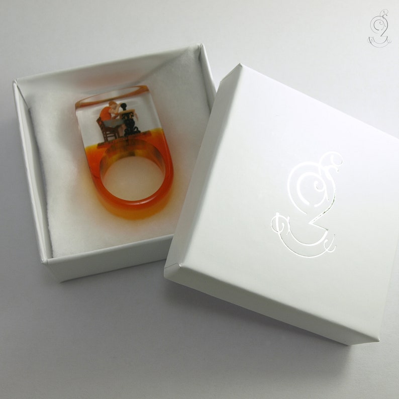 Op de afbeelding: Harsring met een miniatuursc&egrave;ne van een persoon en een hond aan een tafel. De ring heeft een oranje en gele gradi&euml;ntband en wordt gepresenteerd in een witte geschenkdoos.